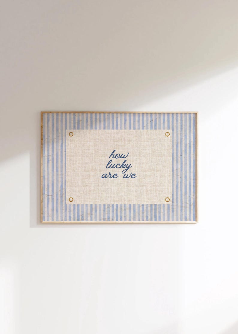 May include: A framed wall art piece with a light blue and white striped background. The center features a beige rectangle with the text "how lucky are we" in blue script. Gold-colored rivets are in each corner.