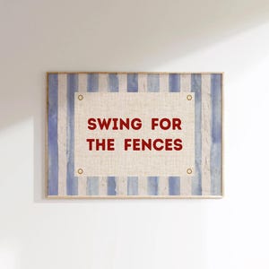Puede incluir: Arte mural enmarcado con un fondo a rayas azules y blancas. El centro presenta un rectángulo beige con las palabras "SWING FOR THE FENCES" en rojo. El marco es de color madera clara.