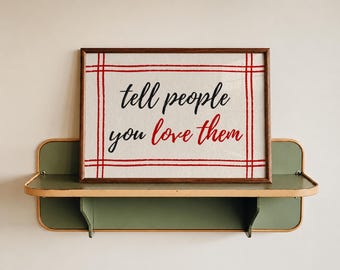 May include: A framed artwork with the message "tell people you love them" in black and red script. The artwork is set against a cream-colored background with a red grid pattern. It rests on a green and tan shelf.