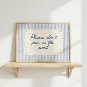 May include: A framed sign with the text "Please don't pee in the pool." The sign has a light blue and white striped border and a beige center. The sign is mounted on a light wood shelf.