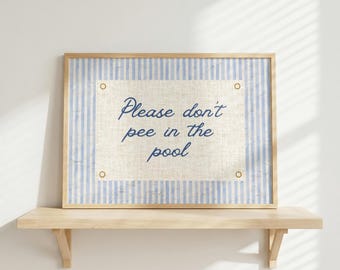 May include: A framed sign with the text "Please don't pee in the pool." The sign has a light blue and white striped border and a beige center. The sign is mounted on a light wood shelf.