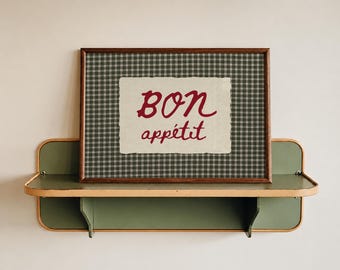 May include: A rectangular framed print featuring a dark green and white checkered pattern with a central cream-colored paper insert displaying the red text "BON appétit" in a handwritten script font, resting on a green metal wall shelf.