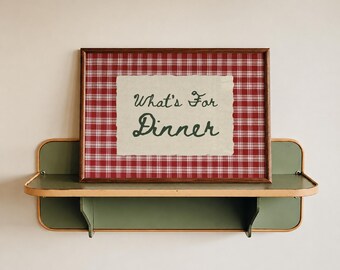 May include: A rectangular framed print featuring a red and white plaid pattern with a central off-white paper cutout displaying the dark green text "What's For Dinner" in a cursive script, resting on a green metal wall shelf with light wood trim.