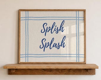 May include: A framed print with the words "Splish Splash" in a dark blue script. The artwork features a light beige background with a blue grid pattern. The frame is made of wood and is displayed on a wooden shelf.