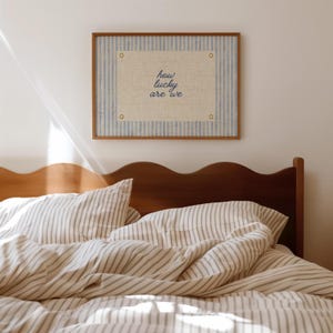 May include: A framed artwork with the text "how lucky are we" hangs above a bed with striped bedding. The artwork features a light blue and white striped background with a central beige rectangle. The bed has a wooden headboard.