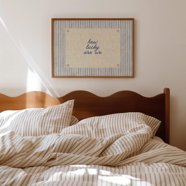 May include: A framed artwork with the text "how lucky are we" hangs above a bed with striped bedding. The artwork features a light blue and white striped background with a central beige rectangle. The bed has a wooden headboard.