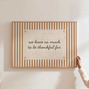 May include: A framed wall art piece with a beige linen center and a striped orange and cream border. The text "we have so much to be thankful for" is printed in dark green. The frame is a light wood color.