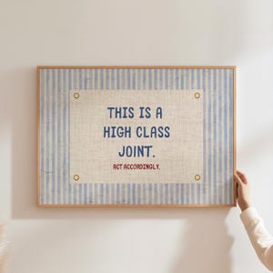 Könnte beinhalten: Ein gerahmter Druck mit einem hellen Holzrahmen. Der Druck hat einen blau-weiß gestreiften Rand und ein zentrales beiges Rechteck mit dem Text "THIS IS A HIGH CLASS JOINT. ACT ACCORDINGLY." in Marineblau und Rot.