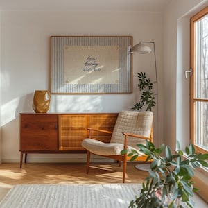 May include: A mid-century modern living room scene featuring a wooden cabinet, a patterned armchair, and a framed artwork with the text "how lucky are we." A gold-colored vase and a floor lamp with a woven shade complete the decor.