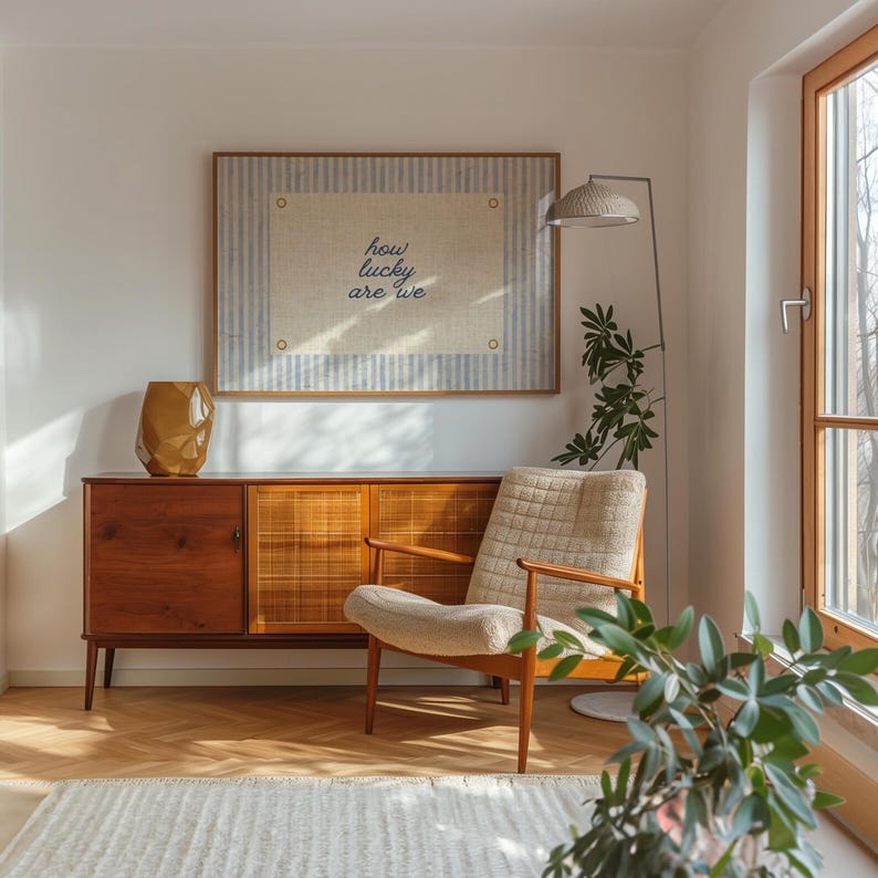 May include: A mid-century modern living room scene featuring a wooden cabinet, a patterned armchair, and a framed artwork with the text "how lucky are we." A gold-colored vase and a floor lamp with a woven shade complete the decor.