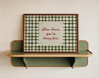 May include: A rectangular wall art print in a dark wood frame featuring a green and white gingham pattern. Centered is a cream-colored square with scalloped edges and four gold grommets, displaying the red text: Slow down, you're doing fine.