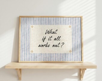 May include: A framed print with the text "What if it all works out?" The print has a light blue and white striped background with a beige center. The frame is a light brown color and the print is displayed on a wooden shelf.