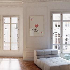 Op de afbeelding: Een witte ingelijste print met een rood hart en de tekst "Feelin' lucky" op een witte achtergrond. De print hangt aan een witte muur in een kamer met een witte bank en een raam.