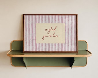May include: A rectangular print in a dark wood frame featuring a light pink and white vertical striped background with a cream-colored center rectangle containing the red cursive text "so glad you're here" resting on a green metal wall shelf.