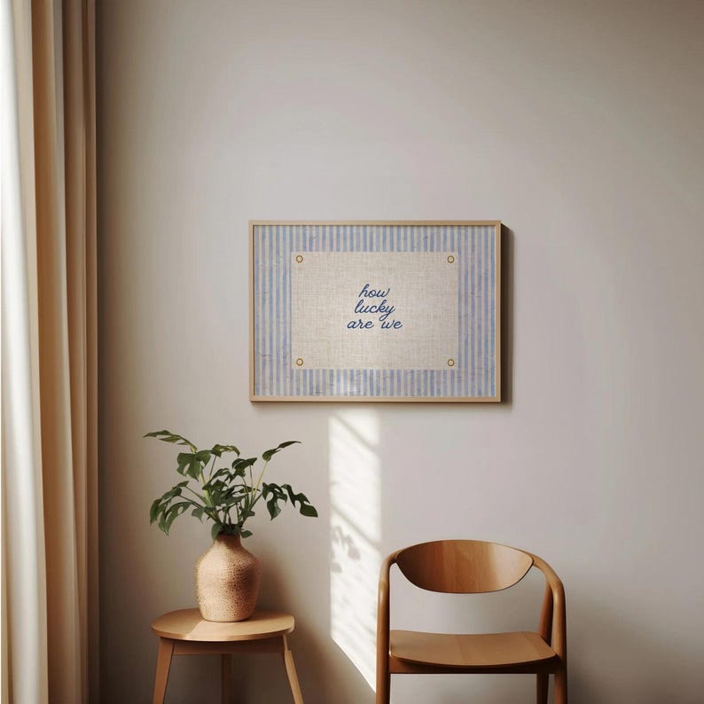 May include: A framed artwork with the text "how lucky are we" in blue script. The artwork features a light-colored linen-like background with a blue striped border. The frame is made of light-colored wood. The artwork is hanging on a neutral-colored wall.
