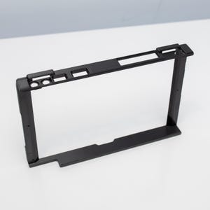 May include: A black plastic frame with rectangular and square cutouts. The frame has a textured surface on one side and is designed with several small holes and a raised section on the top edge. The frame is set against a white background.