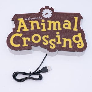 Pu&ograve; includere: Un cartello marrone e giallo "Welcome to Animal Crossing" con una grafica a orologio. Il cartello ha un cavo USB collegato. Il testo &egrave; giallo con sfondo marrone e bordo bianco.