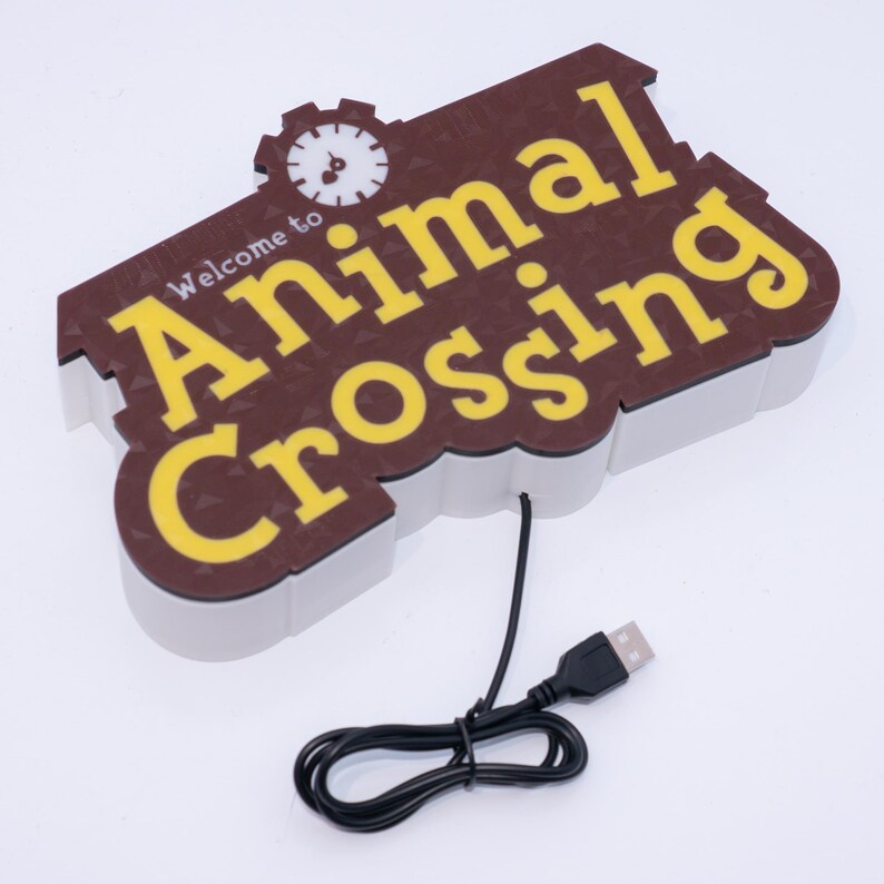 Pu&ograve; includere: Un cartello marrone e giallo con la scritta "Welcome to Animal Crossing" e una grafica a orologio. Il cartello ha un bordo bianco e un cavo USB nero collegato.