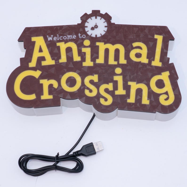 Pu&ograve; includere: Un cartello marrone e giallo con la scritta "Welcome to Animal Crossing" e una grafica a orologio. Il cartello ha un cavo USB collegato per l'alimentazione.