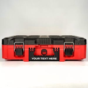 May include: A red and black toolbox with a rectangular shape. The toolbox has a black top with a textured design and a red base. It features two metal latches and the text "YOUR TEXT HERE" on the front.