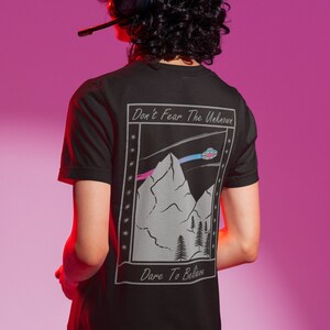 Könnte beinhalten: Schwarzes T-Shirt mit einem Grafikdruck von Bergen, einer fliegenden Untertasse und dem Text "Don't Fear The Unknown" und "Dare To Believe".