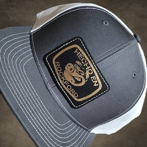 May include: Gray and white baseball cap with a black leather patch that says "Hecho en Rockford" in gold lettering.