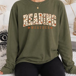 May include: An olive green long-sleeve crewneck sweatshirt featuring the text READING WEATHER in a white block font filled with autumn-themed illustrations of orange pumpkins, brown acorns, and multicolored fall leaves.