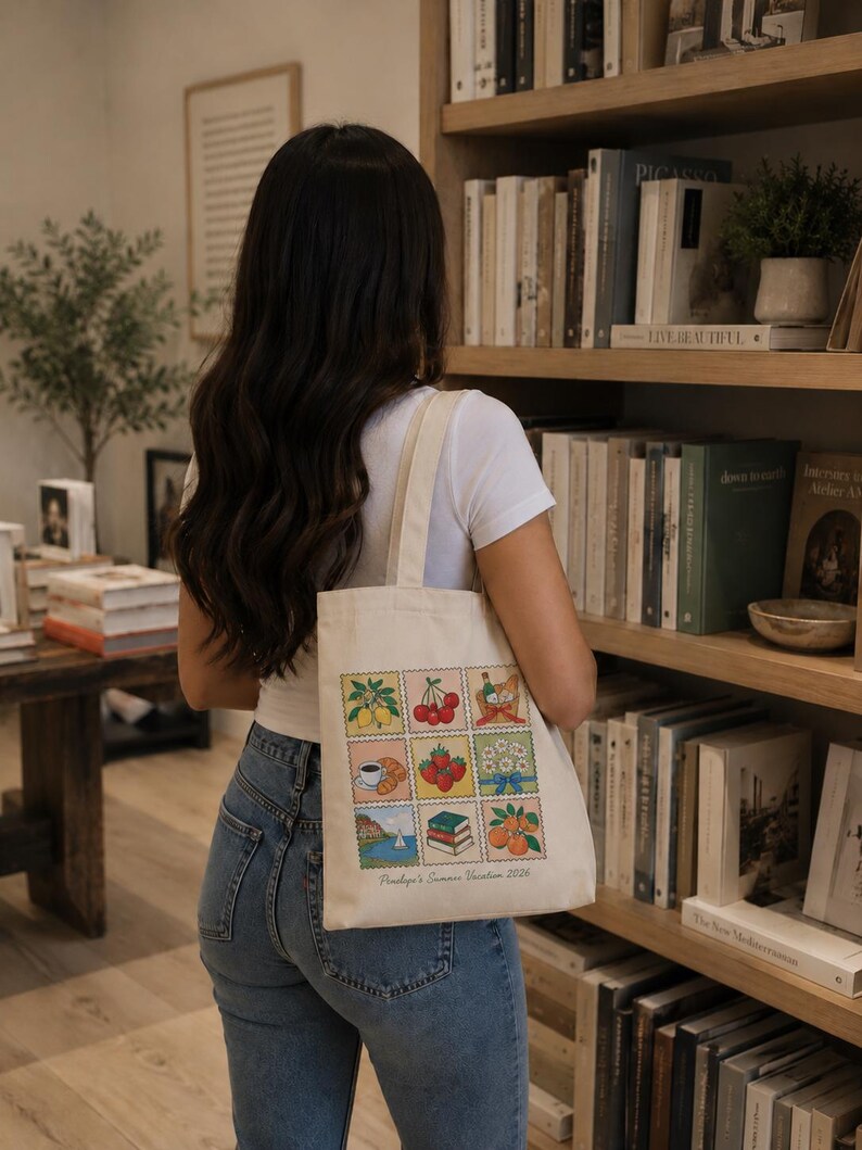 May include: A cream-colored canvas tote bag featuring a grid of nine colorful illustrations resembling postage stamps, including lemons, cherries, a picnic basket, coffee, strawberries, flowers, a coastal scene, books, and oranges, with the text Penelope's Summer Vacation 2026.
