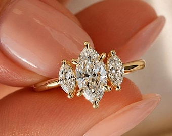 May include: A gold ring featuring a central marquise-cut diamond flanked by two smaller marquise-cut diamonds. The stones are set in a prong setting on a thin, polished gold band, creating a three-stone design with clear, faceted gems.