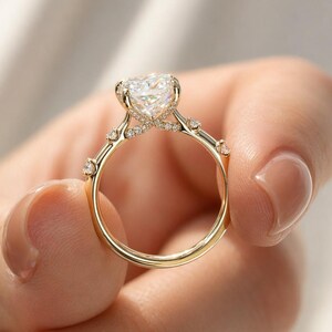 May include: A gold engagement ring featuring a heart-shaped diamond. The band is embellished with smaller diamonds and delicate detailing. The ring is held up by a person's fingers, highlighting its design and craftsmanship.