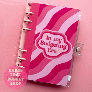 Puede incluir: Una agenda de rayas rosa y blancas con una pegatina que dice "In my Budgeting Era". La agenda tiene una cubierta de plástico transparente y un sistema de encuadernación en espiral.