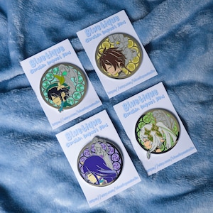 Könnte beinhalten: Vier Emaille-Pins mit Charakteren aus dem Videospiel Genshin Impact. Jeder Pin ist rund und zeigt einen anderen Charakter mit einem Buntglas-Design. Die Pins befinden sich auf weißen Karten mit dem Text "Bluetique Genshin Impact Pins" und einer Website-Adresse.
