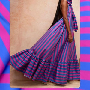 May include: A woman wearing a wide-brimmed hat and a long, striped dress with a ruffled hem. The dress is a mix of pink and blue stripes. The woman is standing in front of a wall.