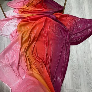 Peut inclure: Un tissu chatoyant aux couleurs vives, allant du rose à l'orange, puis au rouge et au violet. Le matériau est drapé, créant des plis et des textures, avec une finition réfléchissante et brillante.