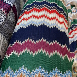 Puede incluir: Primer plano de un rollo de tela con un colorido patrón de zigzag. El diseño presenta bandas alternas de verde, rojo, azul, marrón y morado. La tela tiene una apariencia texturizada, lo que sugiere una construcción tejida o de punto.
