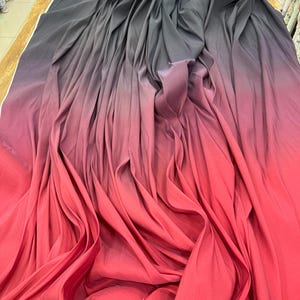 May include: A draped fabric with a gradient effect, transitioning from dark gray at the top to a vibrant red at the bottom. The fabric is soft and flowing, with visible folds and shadows that enhance the color variations. The material appears to be lightweight and suitable for apparel.