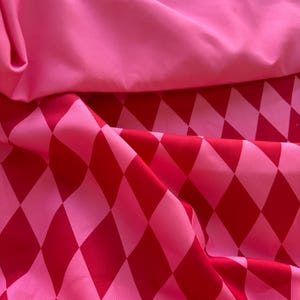 May include: Close-up of pink and red fabric. The top layer is a solid, smooth pink. The bottom layer features a pattern of red diamonds on a pink background. The fabric is folded and draped, creating shadows and texture.