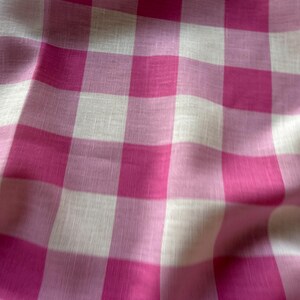Könnte beinhalten: Ein Stoff mit einem klassischen Vichy-Karo-Muster in leuchtenden Magenta-Pink- und klaren Weißtönen. Das Material weist eine sichtbare Webstruktur mit weichen Falten und Drapierungen auf, die ein sich wiederholendes Gitterdesign erzeugen.