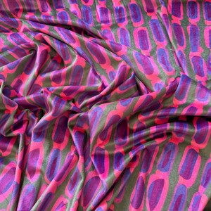 Puede incluir: Una tela con un patrón repetitivo de formas ovaladas alargadas en tonos vibrantes de rosa intenso y morado profundo, sobre un fondo verde oliva oscuro. El material tiene una textura suave similar al terciopelo con pliegues visibles.