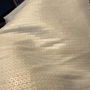 Könnte beinhalten: Nahaufnahme eines hellgrauen Stoffes mit einem sich wiederholenden Muster aus kleinen, glänzenden, kreisförmigen Pailletten. Die Pailletten sind in einem gitterartigen Muster angeordnet und erzeugen eine strukturierte Oberfläche. Der Stoff wirkt weich und fließend.