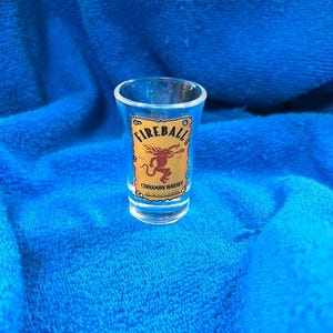 May include: A clear shot glass with the "Fireball Cinnamon Whisky" logo. The logo features the word "Fireball" in a circular design above a red dragon illustration, all on a tan background.