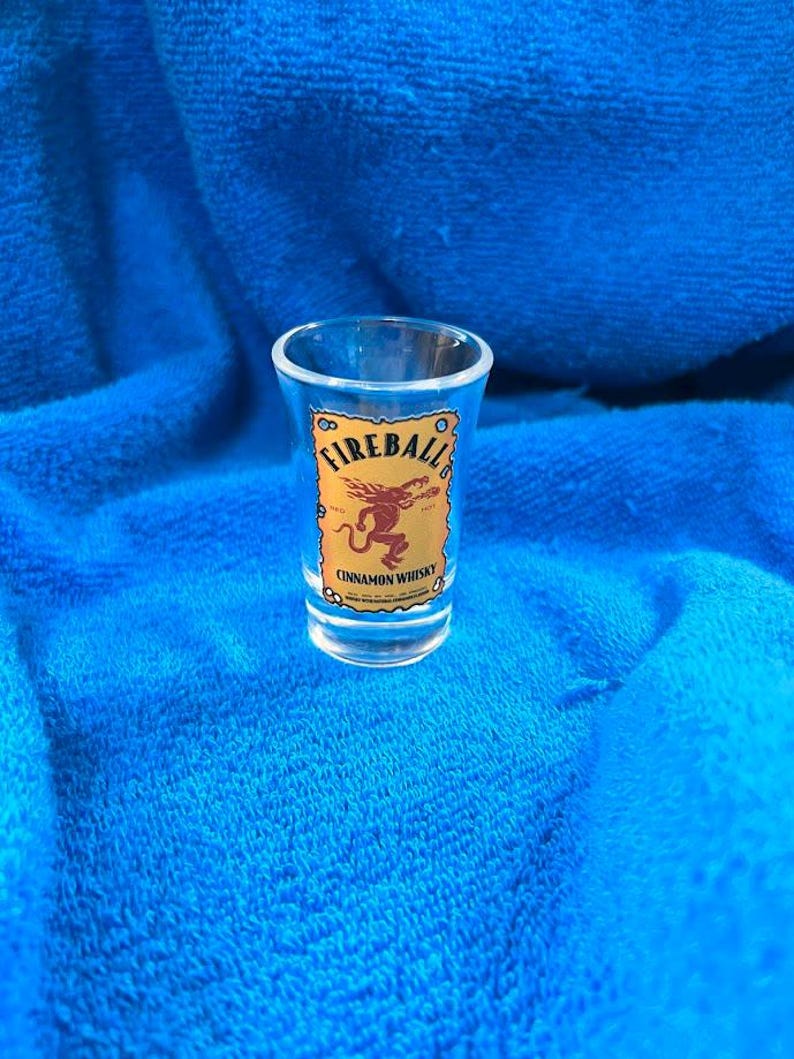 May include: A clear shot glass with the "Fireball Cinnamon Whisky" logo. The logo features the word "Fireball" in a circular design above a red dragon illustration, all on a tan background.