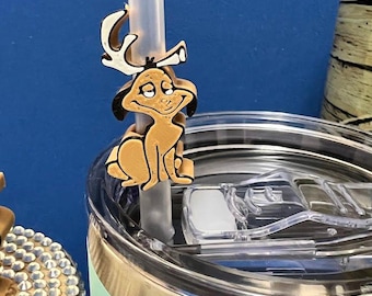 May include: A tan and white cartoon character straw charm is attached to a clear straw. The charm is sitting on the rim of a light green tumbler. The background is a solid blue.