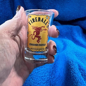 May include: A clear shot glass with the Fireball Cinnamon Whisky logo. The label is yellow with red text and a red cartoon image of a devil. The shot glass is held against a blue background.