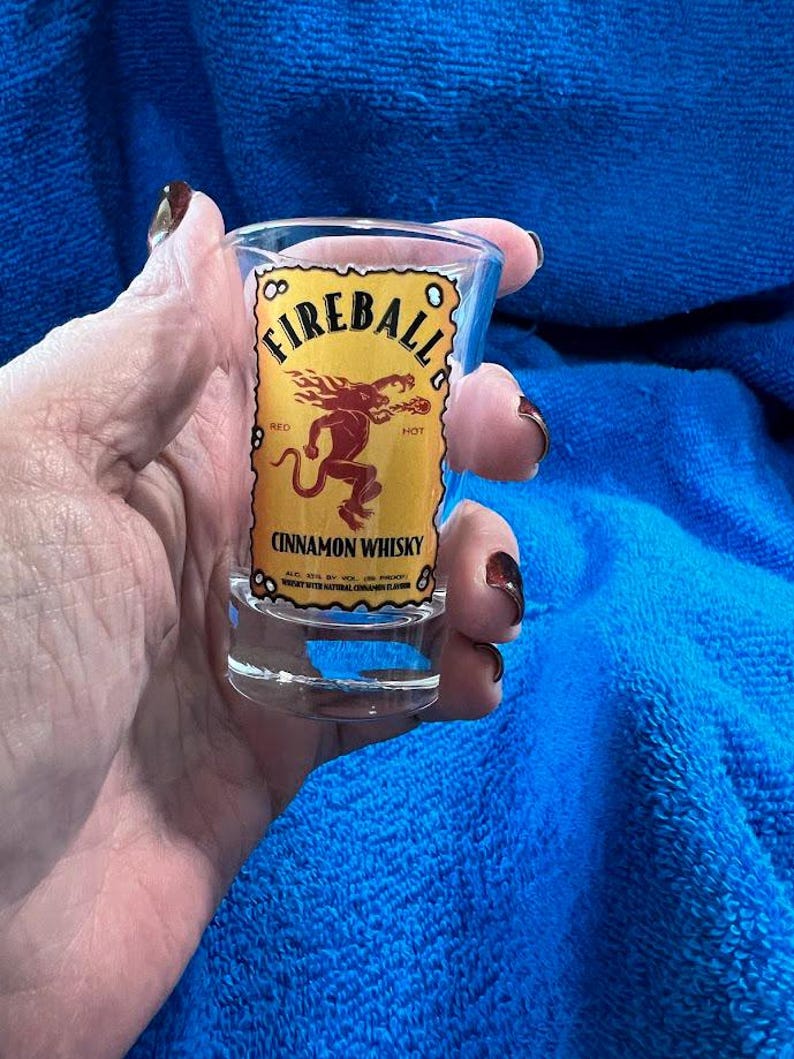 May include: A clear shot glass with the Fireball Cinnamon Whisky logo. The label is yellow with red text and a red cartoon image of a devil. The shot glass is held against a blue background.