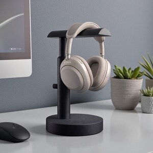 May include: A black headphone stand with a pair of beige headphones. The stand has a circular base and a vertical post with a curved top to hold the headphones. A computer mouse and potted plants are also visible on the white desk.