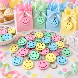 May include: A collection of round smiley face keychains in pastel pink, yellow, blue, and green, scattered alongside four matching gift bags with ribbon handles and a white bowl filled with additional keychains and a polka dot gift box.