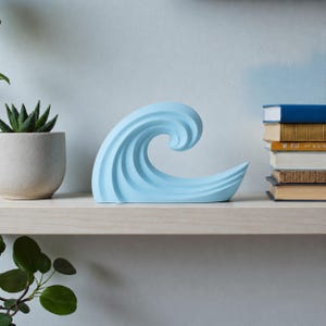 Könnte beinhalten: Ein hellblaues, wellenförmiges Dekorationsstück. Die Skulptur steht auf einem Holzregal, neben einer Topf-Sukkulente und einem Stapel Bücher. Das Wellendesign hat eine geschwungene, fließende Form mit detaillierten Rippen.