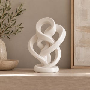 May include: A matte white ceramic sculpture featuring an abstract, continuous looped design with multiple intertwined ribbons that form a complex, open-work structure. The object stands on a circular base and has a smooth, uniform finish throughout.