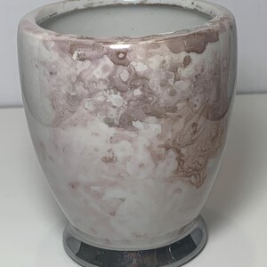 May include: A small, ceramic vase with a marbled design in shades of white, pink, and brown. The vase has a rounded body and a silver-colored base. The interior is a light gray color.
